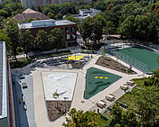 Foto des Forum am Campus Efeuweg, einer multifunktionalen, modernen Landschaftsarchitektur mit Amphitheater, Sportfeld, Gemeinschaftsgarten und Bewegungsflächen. Der Platz ist umgeben von Grünflächen, bietet barrierefreien Zugang und dient als Treffpunkt für Schüler, Nachbarschaft und Veranstaltungen. Das Amphitheater ist mit Sonnensegeln ausgestattet, die Bewegungsflächen sind leicht modelliert, und das gesamte Areal verbindet den Schulhof mit dem öffentlichen Raum in einem urbanen Design.