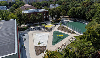 Foto des Forum am Campus Efeuweg, einer multifunktionalen, modernen Landschaftsarchitektur mit Amphitheater, Sportfeld, Gemeinschaftsgarten und Bewegungsflächen. Der Platz ist umgeben von Grünflächen, bietet barrierefreien Zugang und dient als Treffpunkt für Schüler, Nachbarschaft und Veranstaltungen. Das Amphitheater ist mit Sonnensegeln ausgestattet, die Bewegungsflächen sind leicht modelliert, und das gesamte Areal verbindet den Schulhof mit dem öffentlichen Raum in einem urbanen Design.